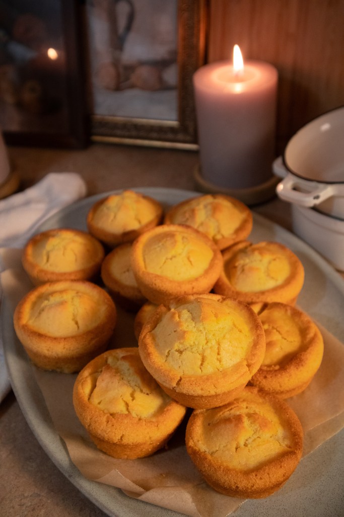 A plate of freshly baked vegan cornbread muffins, placed on a table with a softly glowing candle in the background.