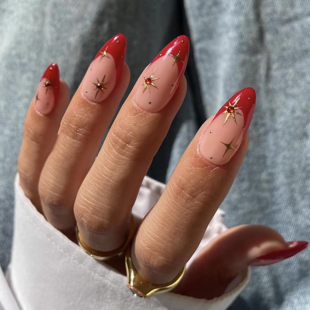 Close-up of a hand with long, pointed nails featuring red tips, nude base, and gold star embellishments.