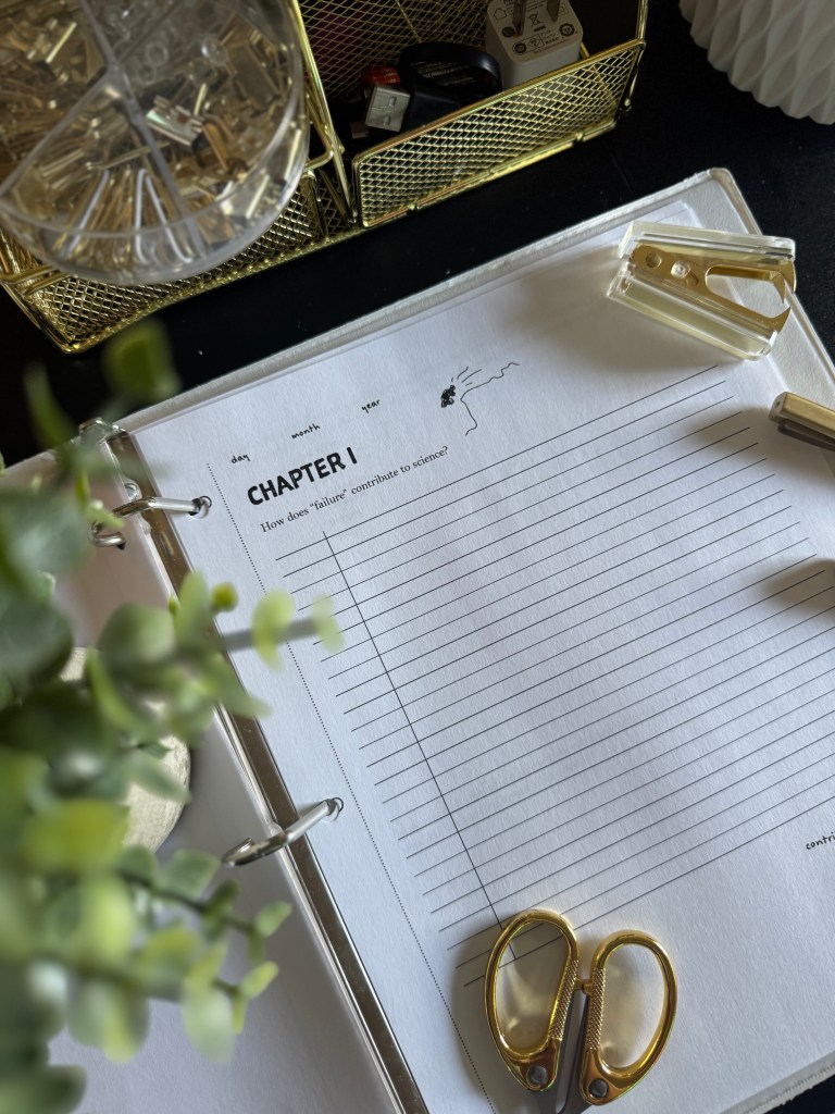 An open notebook page titled 'CHAPTER I' with a question about failure in science, surrounded by stationery items like scissors, a stapler, and a gold storage basket with small items, along with a small green plant.