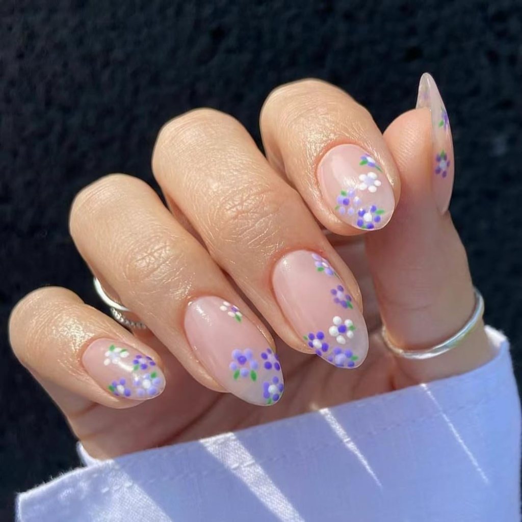 A close-up of a hand displaying manicured nails with a clear polish and delicate floral designs in purple, white, and green.