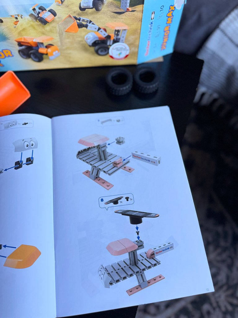 An instructional manual page showing steps to build a construction vehicle with orange and gray parts, alongside a box and two black wheels.