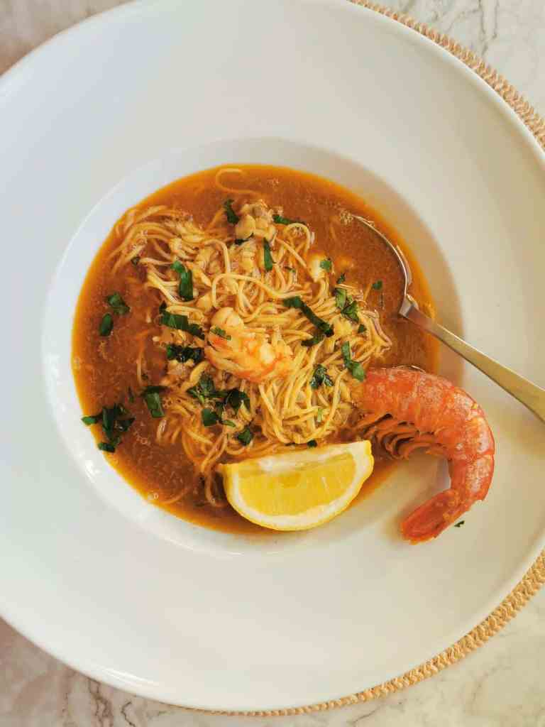 A bowl of shrimp ramen noodle soup garnished with green herbs and a lemon wedge, served on a round white plate.