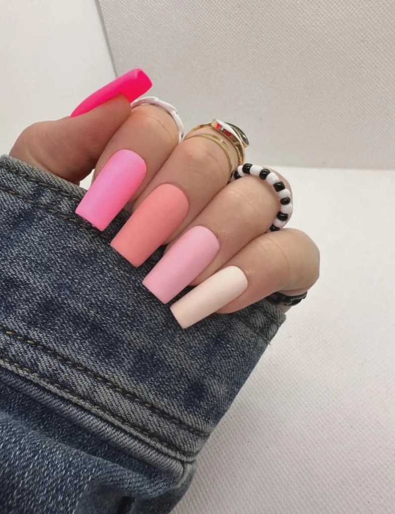 A close-up of a hand with long, brightly colored nails in shades of pink and pastel, displaying various rings, resting on a denim surface.