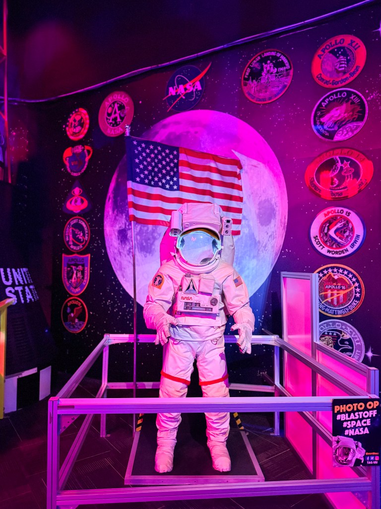 An astronaut mannequin wearing a space suit stands inside a display with a backdrop of the moon and NASA mission patches, alongside an American flag.