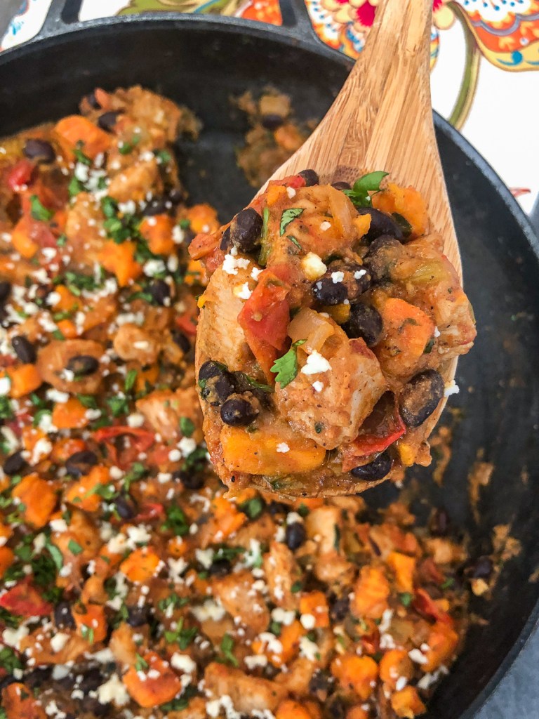 A wooden spoon showcasing a serving of chicken mixed with black beans, sweet potatoes, and colorful diced vegetables, garnished with fresh herbs, served in a cast iron skillet.