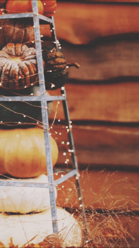 A rustic metal rack stacked with various pumpkins, adorned with fairy lights, set against a warm wooden background, capturing the essence of fall decor.