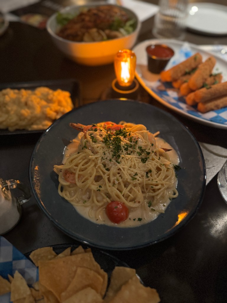 A plate of shrimp fettuccine pasta garnished with cherry tomatoes and parsley, with additional dishes in the background including macaroni and cheese, fried mozzarella sticks, and a salad bowl. Great Wolf Lodge in Grapevine, TX 