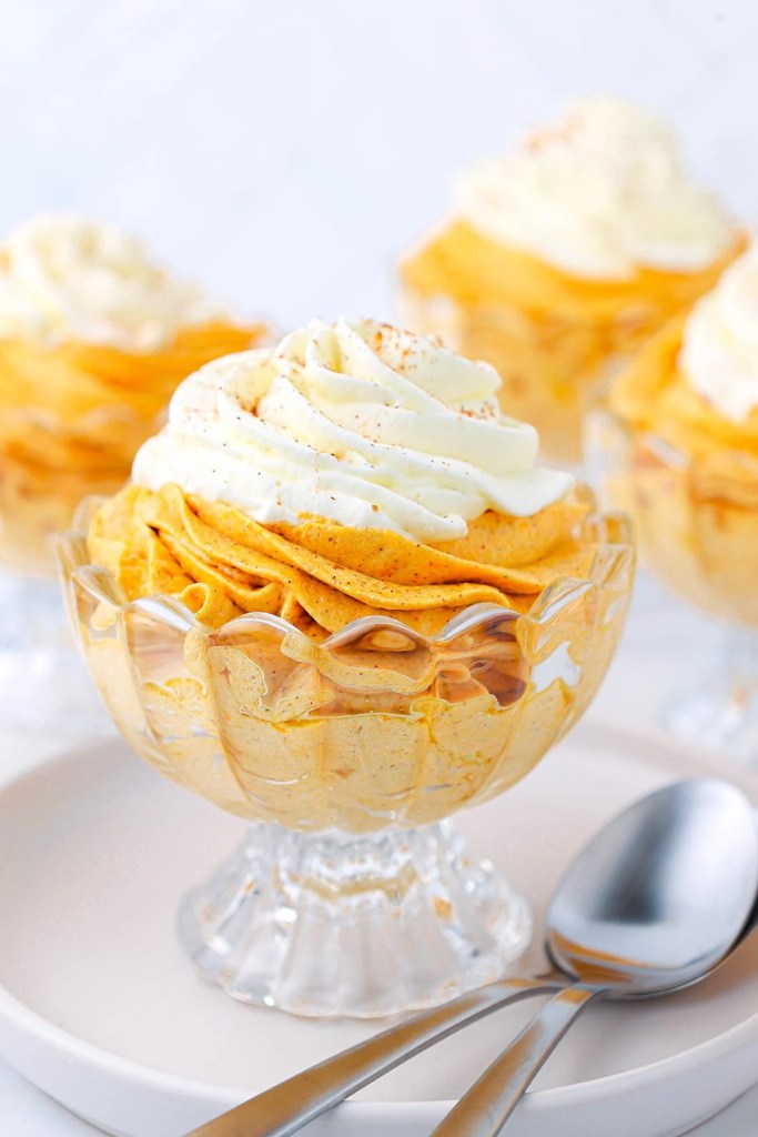 A clear glass dessert cup filled with creamy pumpkin mousse topped with whipped cream, garnished with a sprinkle of spice.