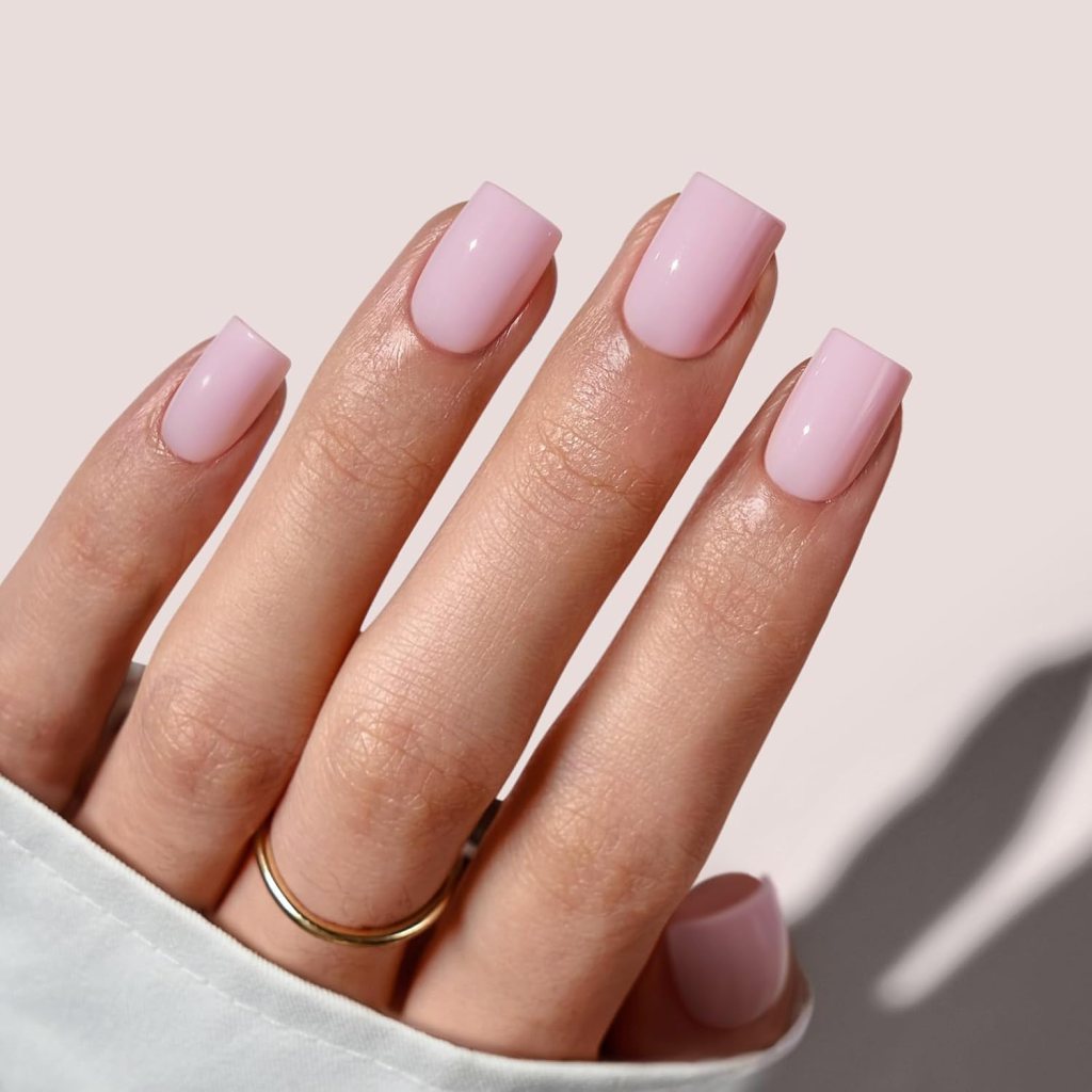 Close-up of a hand with long, square-shaped nails painted in a light pink color, adorned with a thin gold ring.