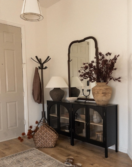 A cozy entryway featuring a black cabinet with glass doors, a large mirror above it, and a decorative vase filled with autumnal foliage. A lamp sits on the cabinet next to a woven basket, with a coat rack visible in the background.