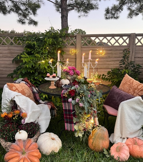 A cozy outdoor fall tablescape featuring a black round table adorned with a floral centerpiece, candles, and pumpkins. Soft blankets and pillows provide comfort, while twinkling lights create a magical atmosphere.