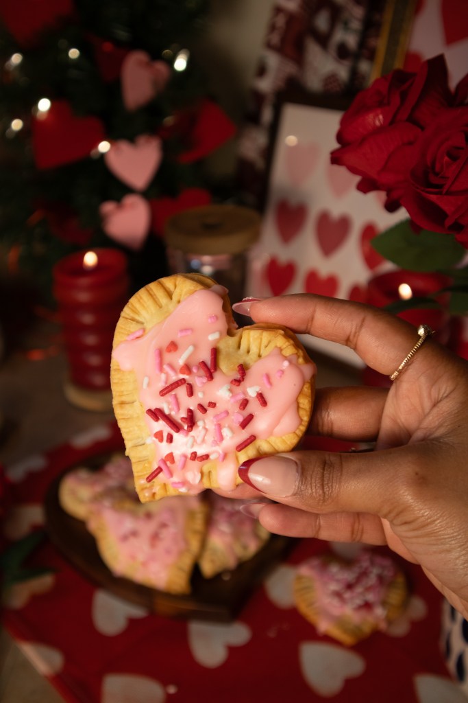 A hand holding a heart-shaped pop tarts topped with pink icing and colorful sprinkles, with more pastries in the background and festive decorations.