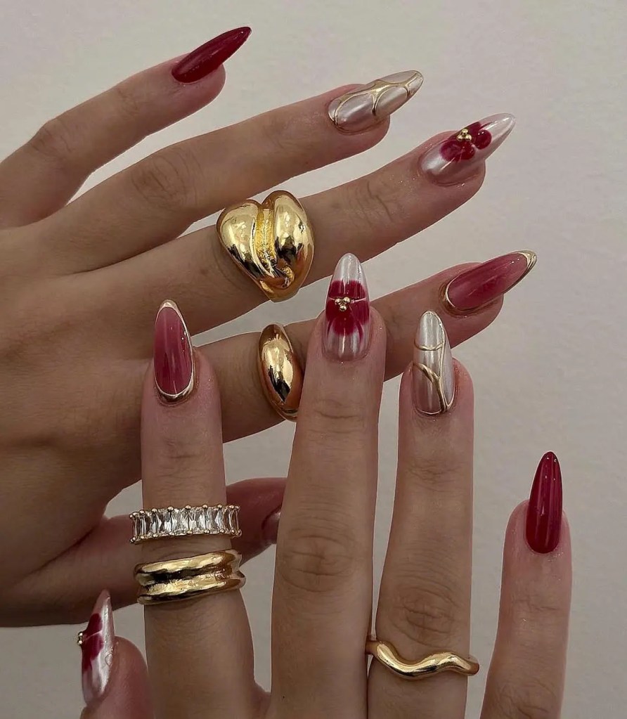 Close-up of hands showcasing stylish manicured nails in various designs, including red, metallic, and floral patterns, adorned with multiple gold rings.