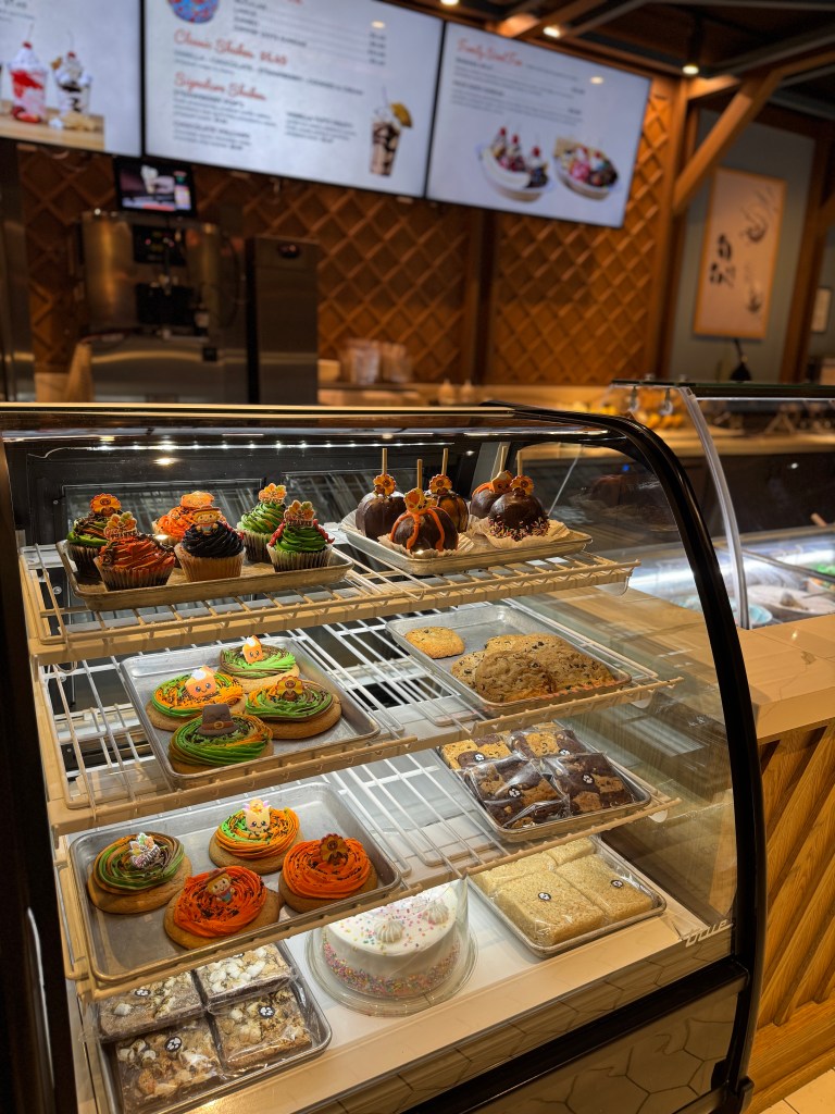 Display case filled with colorful and festive desserts including cupcakes, cookies, and a cake, with a menu board visible in the background.