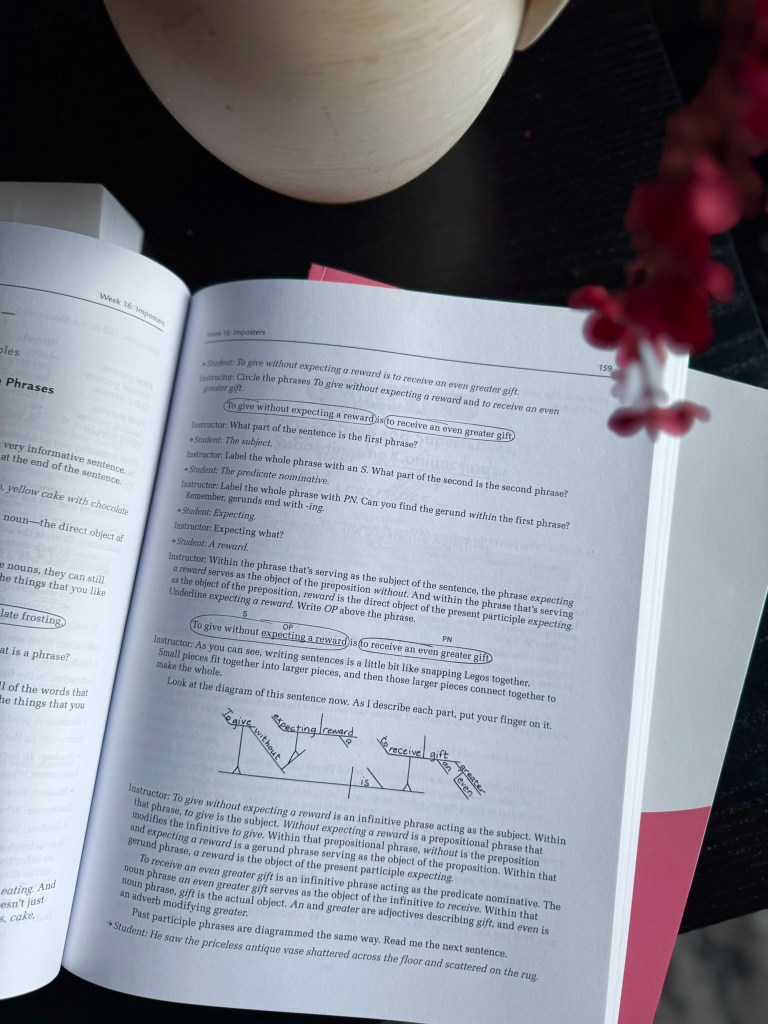A close-up view of an open textbook page with educational content about sentence structure, focusing on phrases and their grammatical roles. A potted plant and a partially visible object can be seen in the background.