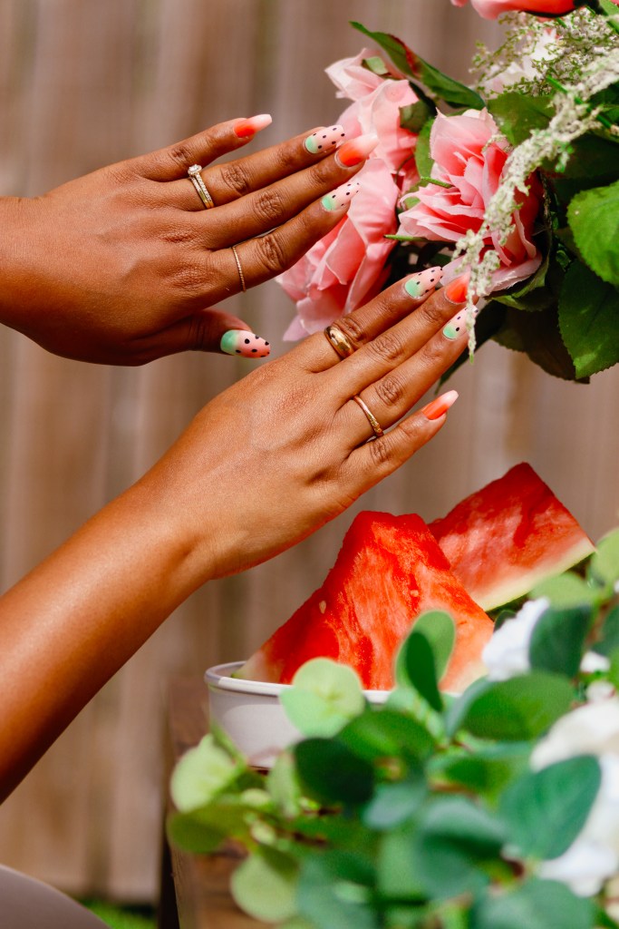 Close-up of two hands with colorful nail art, gently touching pink flowers, with slices of watermelon and greenery in the foreground.