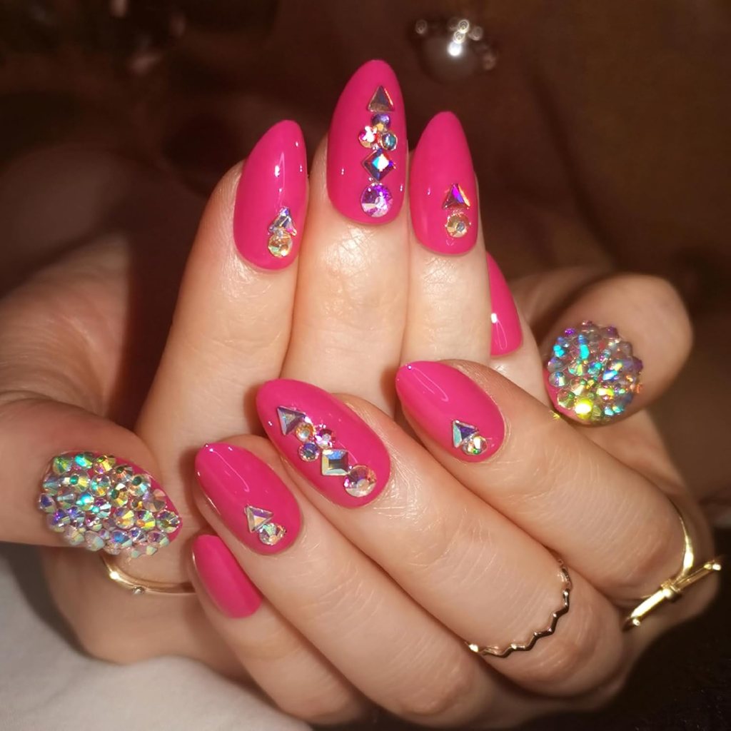 Close-up of hands displaying vibrant pink nails adorned with various colorful gemstones and rhinestones.