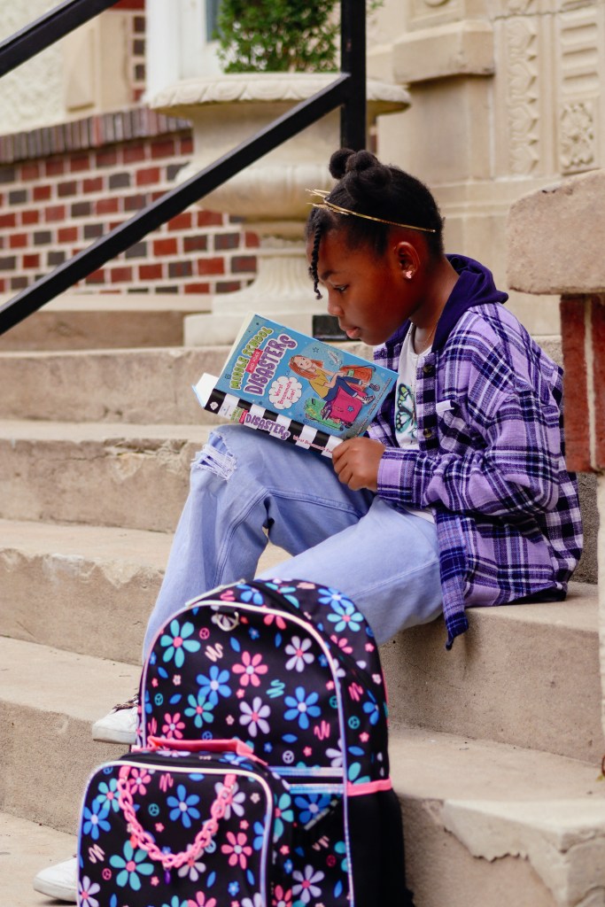 Tween girl reading Middle School and Other Disasters while sitting on outdoor steps with her backpack.