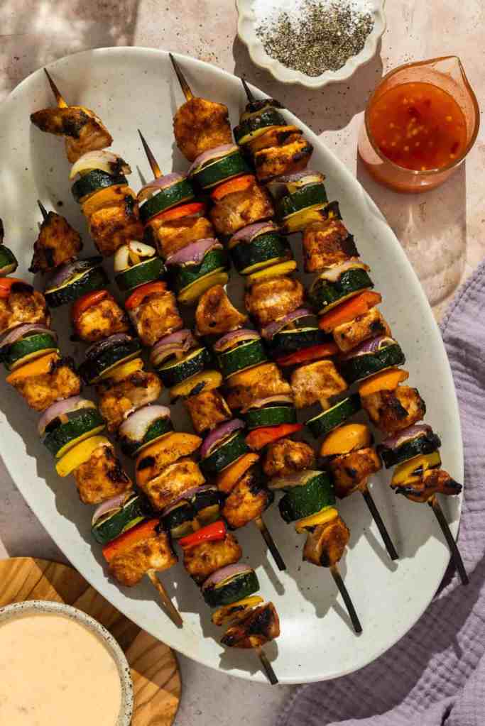 A platter of grilled chicken and vegetable skewers, beautifully arranged with colorful bell peppers, zucchini, and onions, with a side of dipping sauce and seasoning.