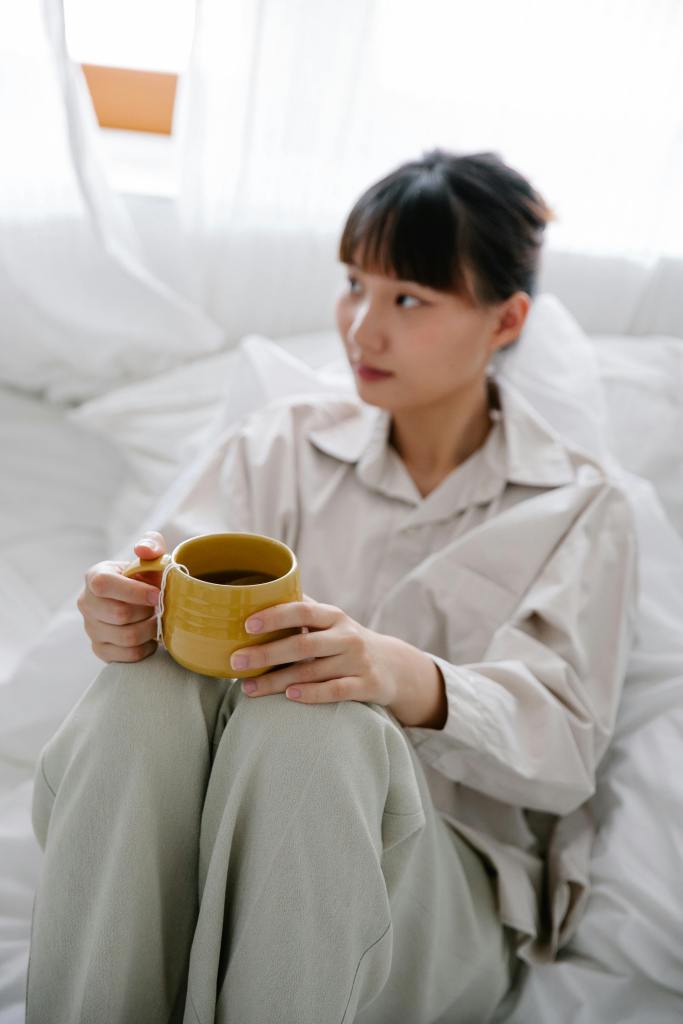 A person sitting on a bed holding a yellow mug, wrapped in cozy clothing, with a serene expression, surrounded by soft bedding and natural light filtering through sheer curtains.