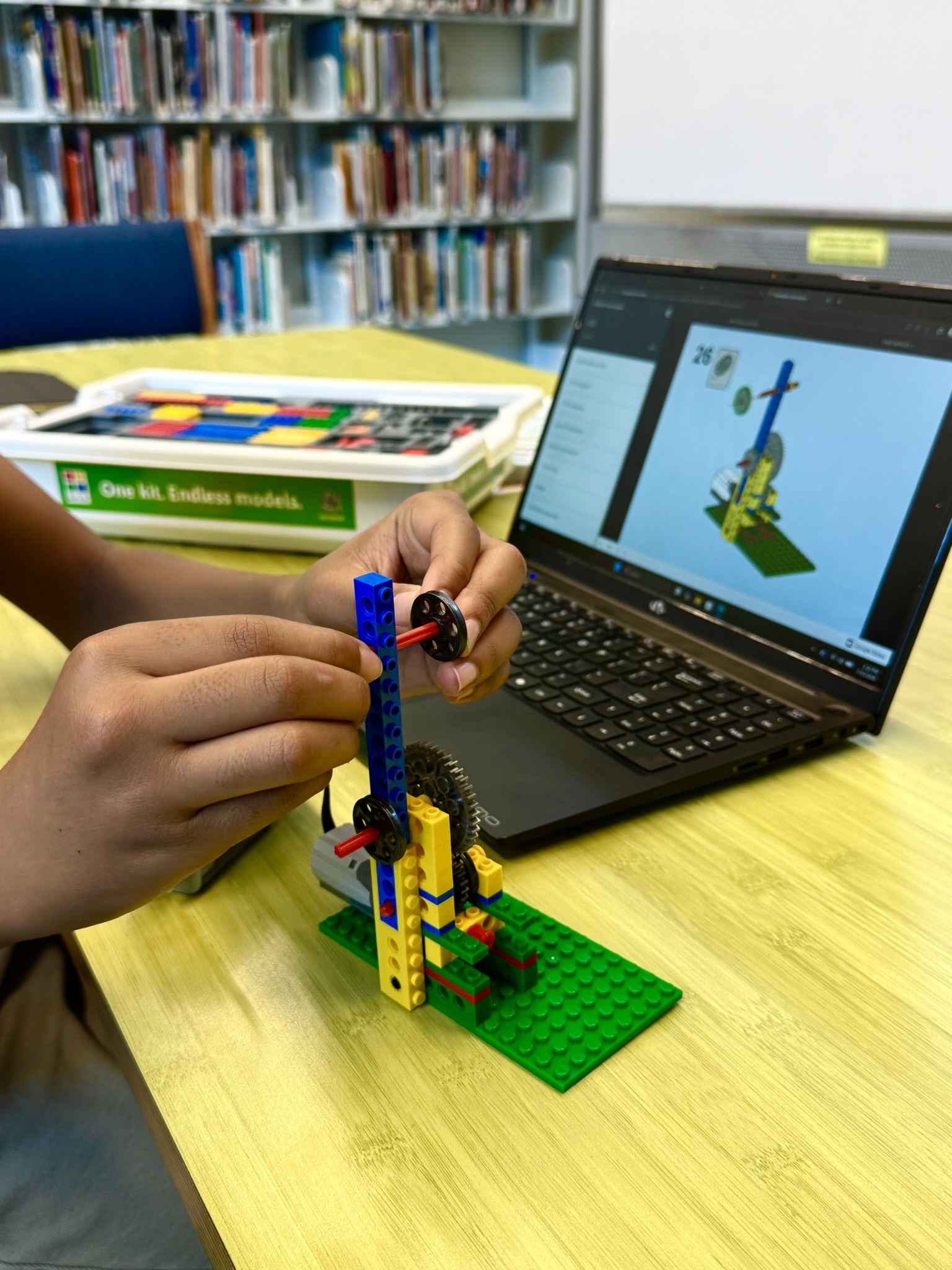 Hands assembling a LEGO structure while following instructions on a laptop screen in a library setting