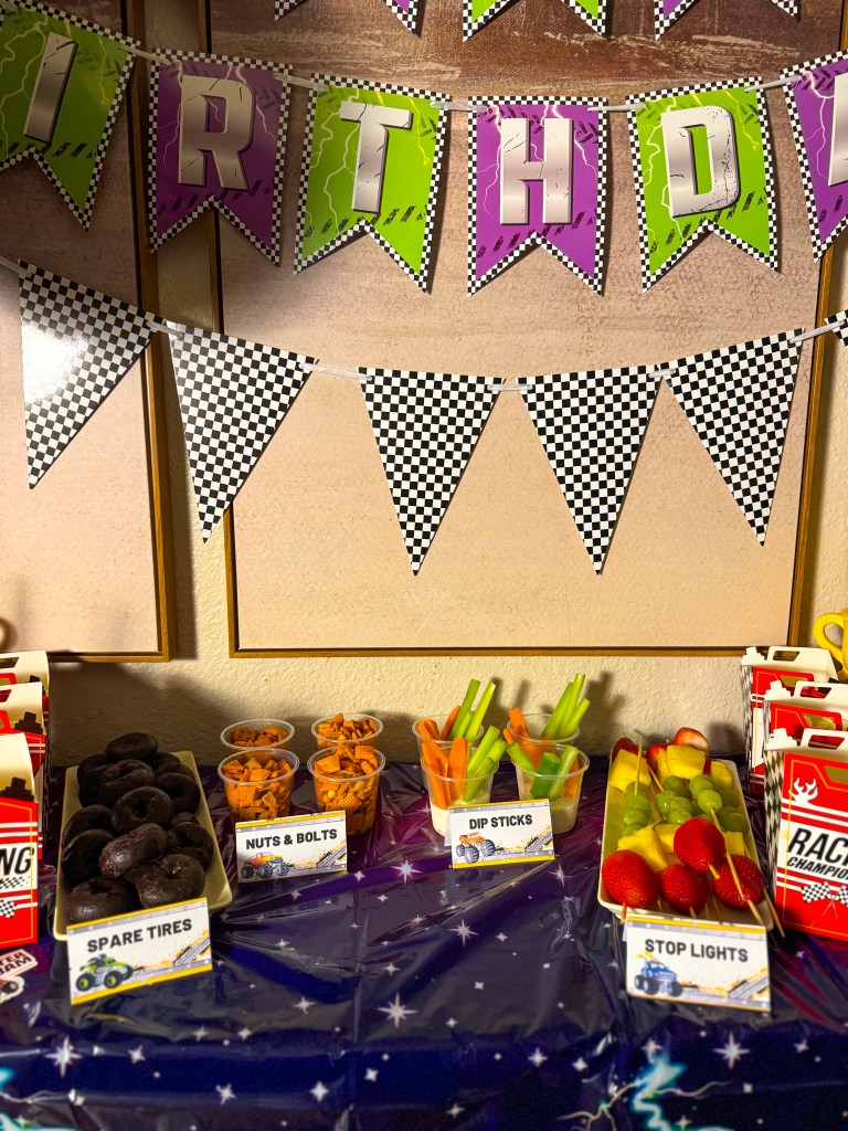 Monster truck birthday party snack table with racing themed decorations, happy birthday banner, and creative snacks like spare tires donuts, nuts and bolts mix, dip sticks vegetables, and stop light fruit skewers.