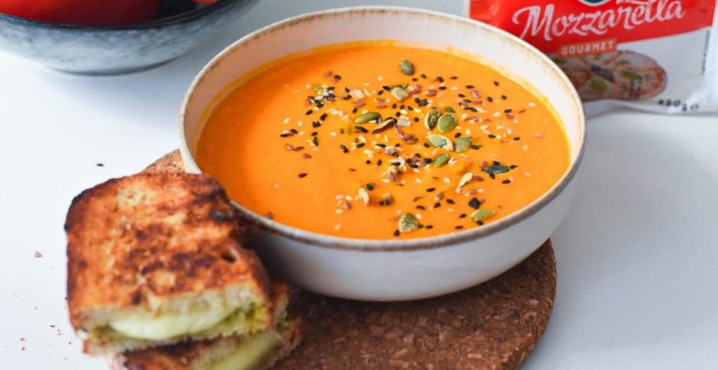 A warm bowl of orange soup topped with seeds and spices, accompanied by a golden grilled cheese sandwich on a cork coaster.