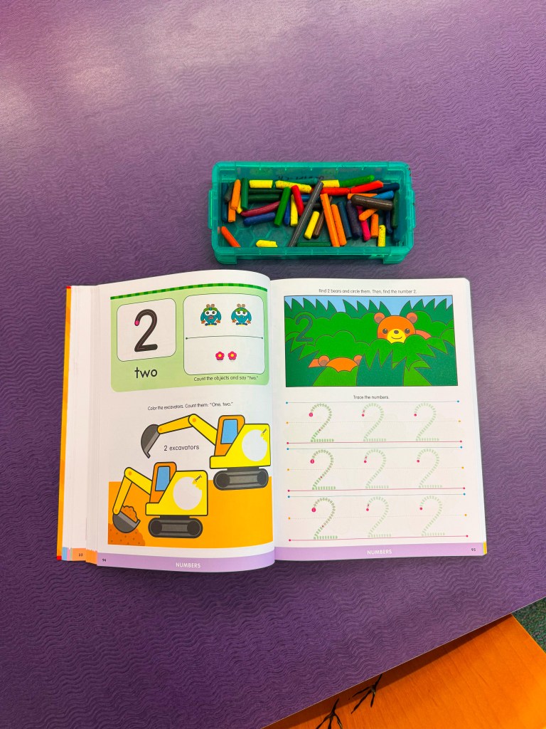 An educational workbook opened to the number two with colorful illustrations and exercises, accompanied by a container of colorful counting sticks on a purple surface.