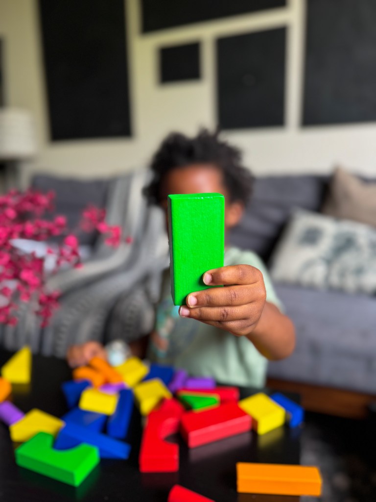 A child holding a green building block in front of a scattered collection of colorful blocks on a table.