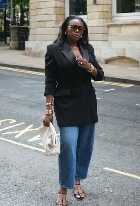 A woman wearing a black oversized blazer, blue wide-leg jeans, and strappy sandals stands on a city street, holding a light-colored handbag.