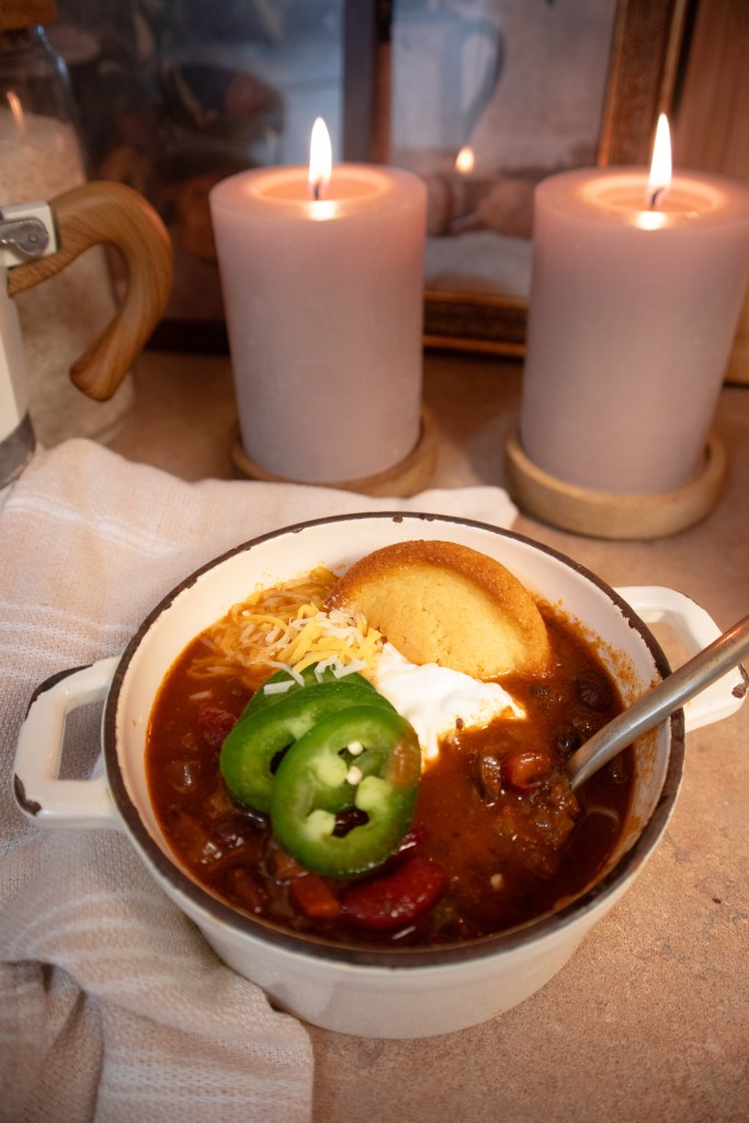 A bowl of vegan mushroom chili topped with sour cream, cheese, jalapeño slices, and a piece of cornbread, placed on a countertop with candles in the background.