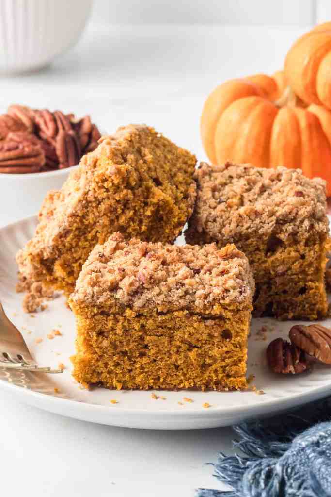 A close-up of pumpkin crumb cake squares topped with a streusel topping, served on a white plate with pecans and a small pumpkin in the background.
