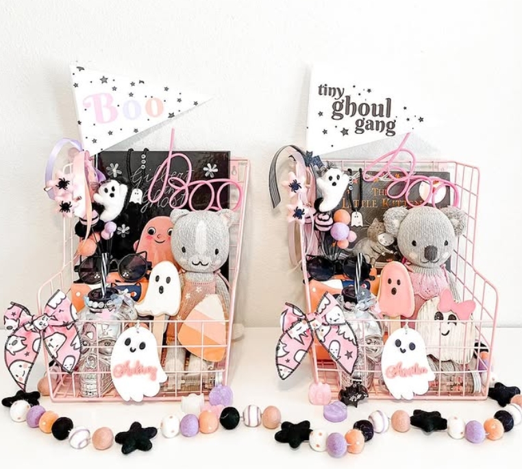 Two pink wire baskets filled with Halloween-themed goodies, including plush toys, cookies, and decorations, with flags reading 'Boo' and 'tiny ghoul gang'.