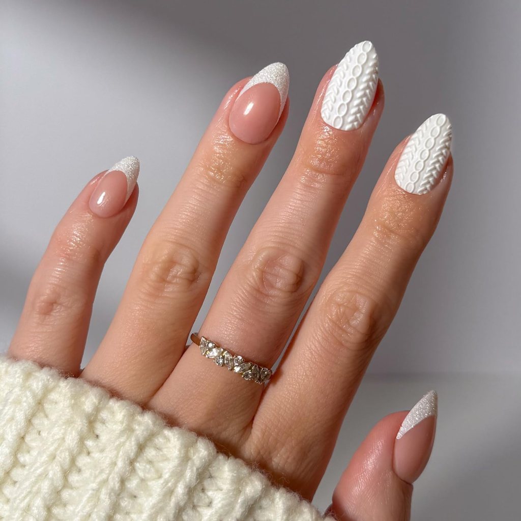 A close-up of a hand with stylish manicure featuring nude and white textured nails, some with a glossy finish and others with a patterned design. A silver ring is worn on one finger, and the hand is resting on a cream knit sweater.