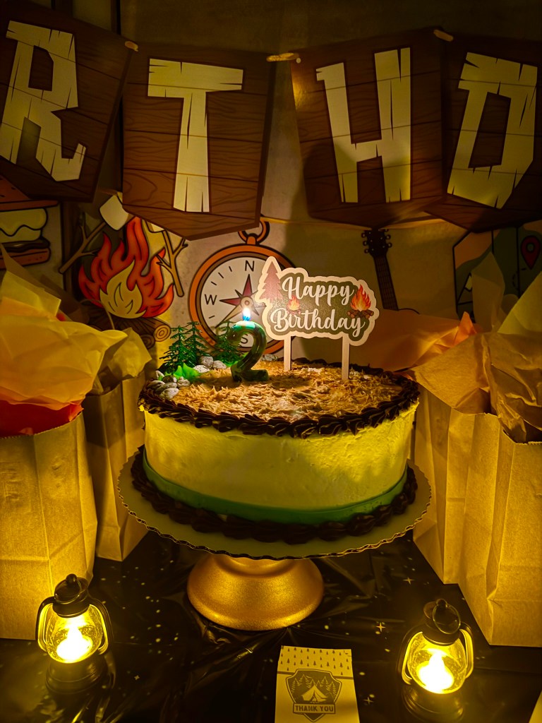 Camping birthday party cake displayed on a gold cake stand with lanterns and favor bags.