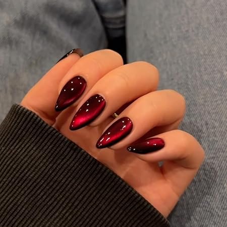 A close-up of a manicured hand displaying deep red, glossy stiletto nails with a striking cat-eye effect, complementing a cozy sweater.