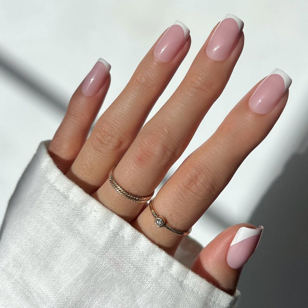 A close-up of a hand with elegantly manicured nails featuring a pink base and white tips, adorned with three delicate rings, partially covered by a light-colored fabric.