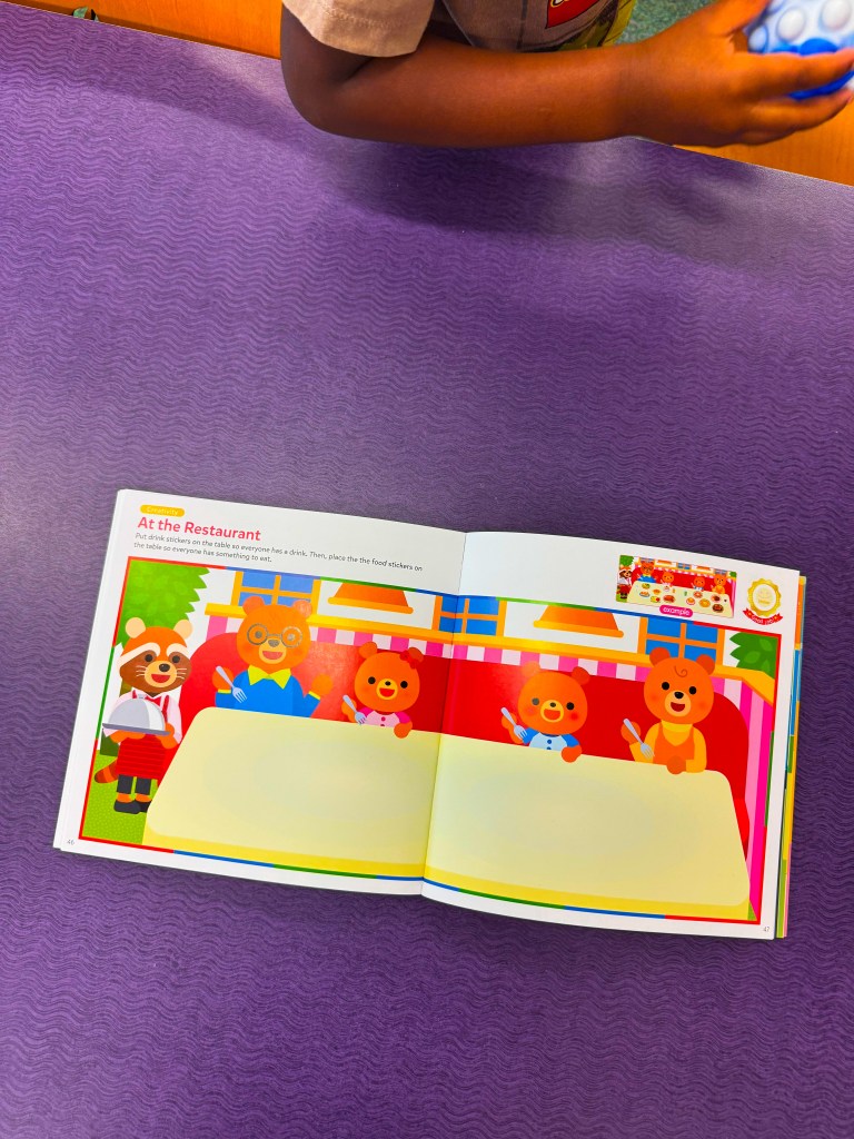 An open children's book titled 'At the Restaurant', featuring cartoon animal characters at a dining table in a colorful restaurant setting. A child's hand is partially visible holding a blue toy.