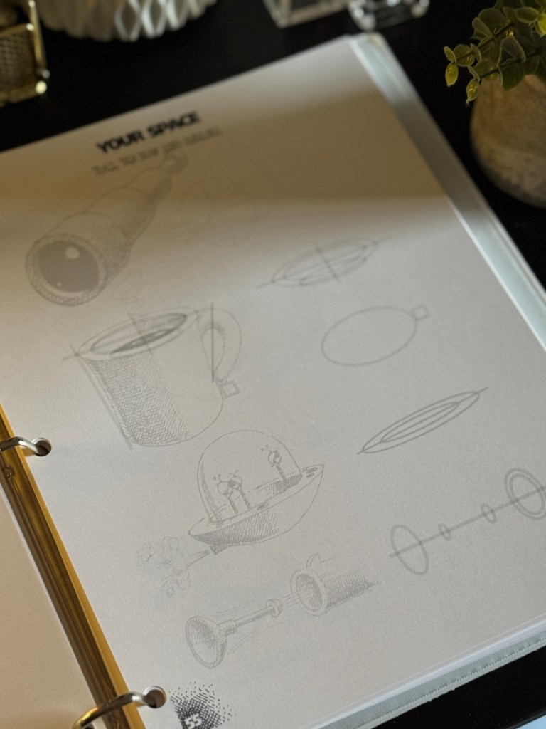 A close-up of a drawing page in a binder, featuring sketches of various objects, including a flashlight, a cup, and a spaceship. The page is titled 'YOUR SPACE'.
