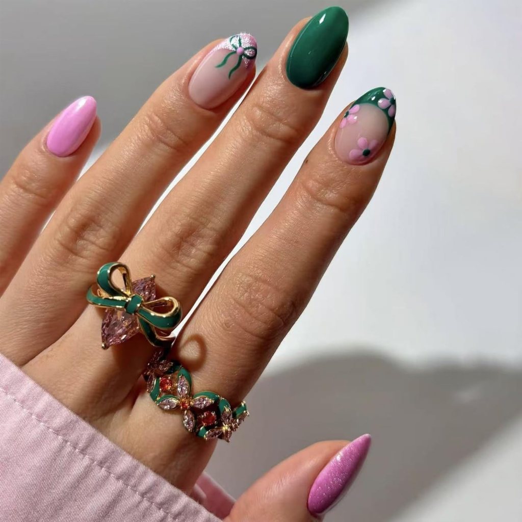 A close-up view of a hand displaying colorful nail art with various designs including green, pink, and floral patterns, adorned with two decorative rings.