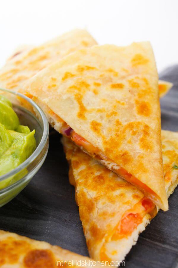 Close-up of cheesy chicken quesadillas on a black plate, accompanied by a small bowl of guacamole.
