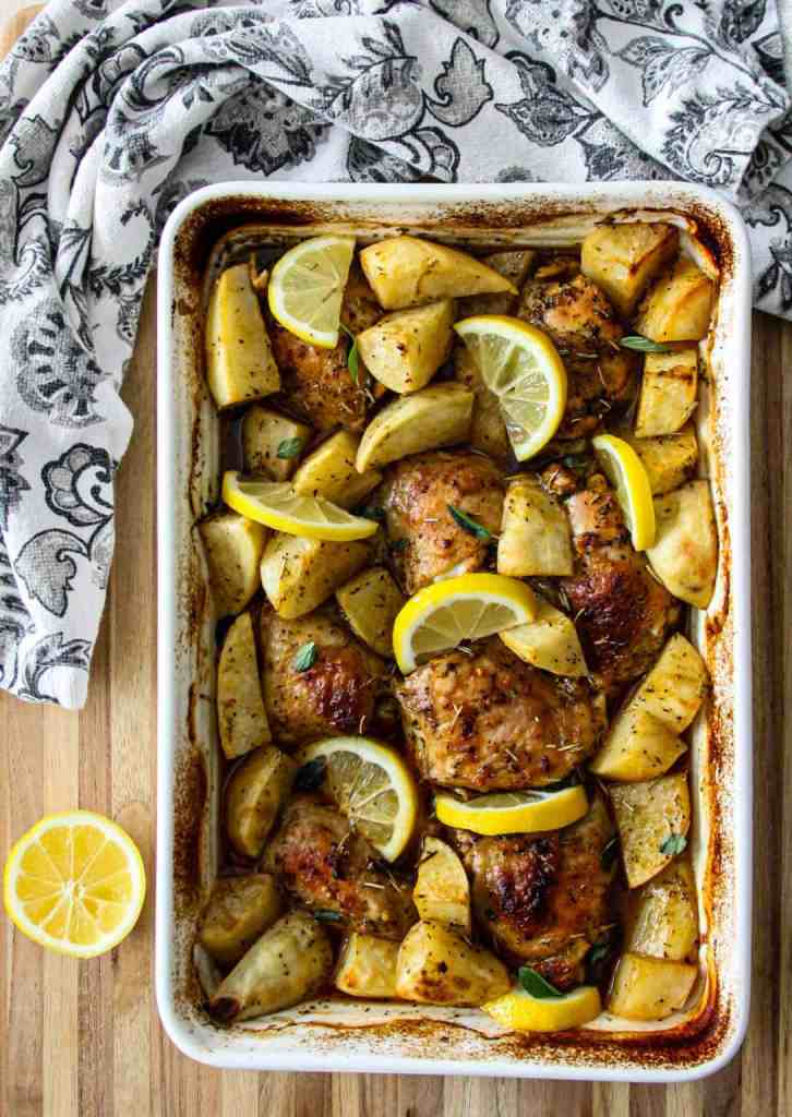 Baked chicken thighs with lemon slices and roasted potatoes in a white baking dish, surrounded by a patterned cloth on a wooden surface.