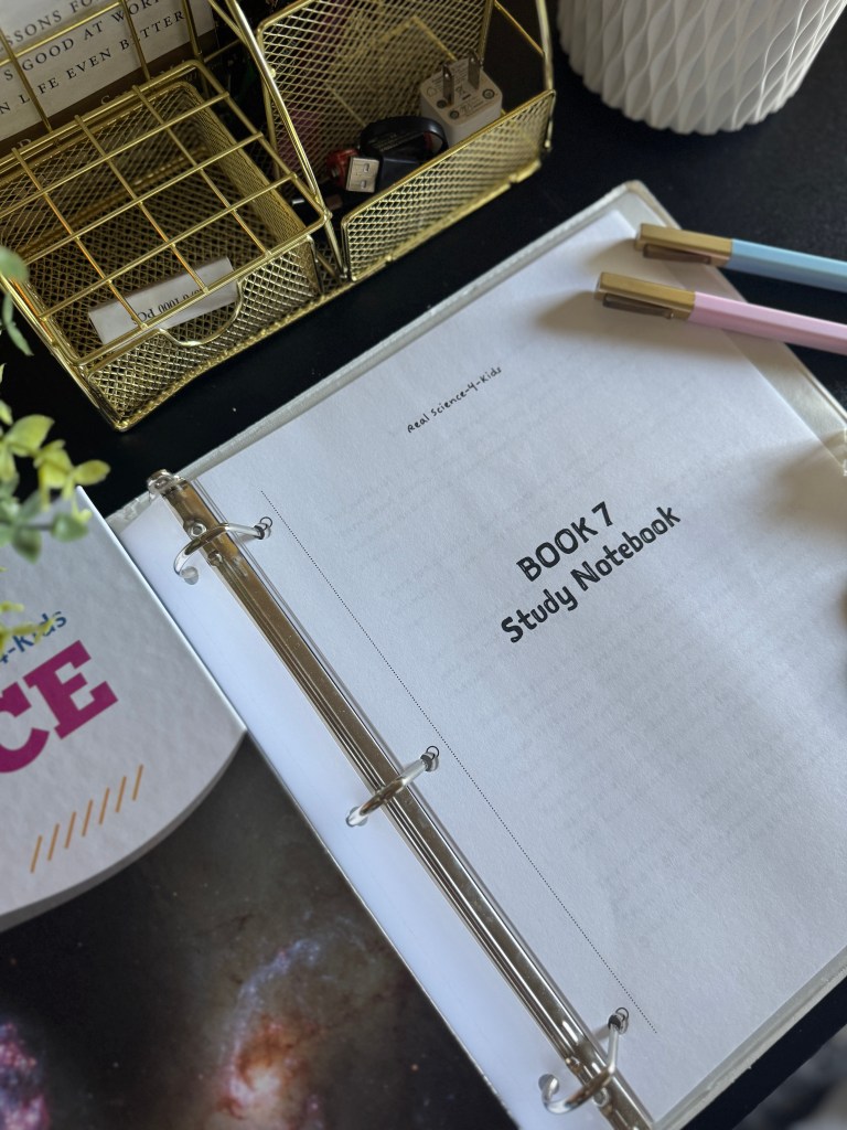 A close-up image of an open binder showcasing the title page of 'Real Science-4-Kids Book 7 Study Notebook'. Surrounding items include gold organizers containing various supplies, a decorative white planter, and a colorful notebook with the word 'SCIENCE' visible.