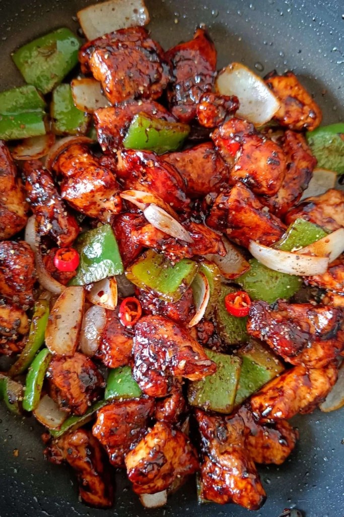 A close-up view of stir-fried chicken pieces mixed with green bell peppers and onions, glazed in a shiny, dark sauce, garnished with red chili slices.