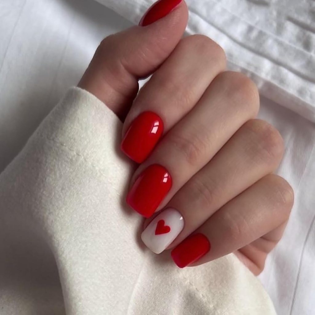A hand with red polished nails, featuring a design of a small red heart on the white nail of the ring finger.