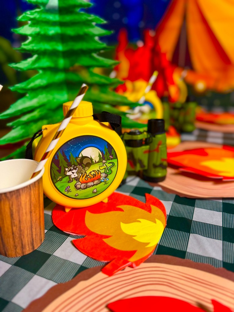 Camping themed birthday party table with yellow canteens, green binoculars, fire napkins, and wooden cups.