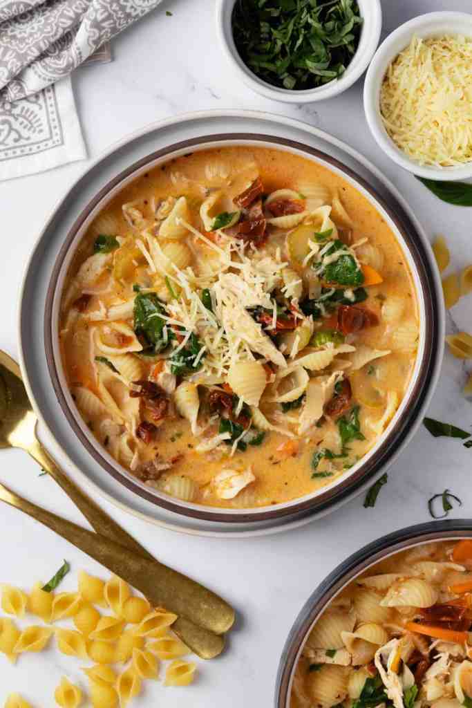 A bowl of creamy chicken pasta soup garnished with herbs and cheese, surrounded by uncooked pasta and additional garnishes.