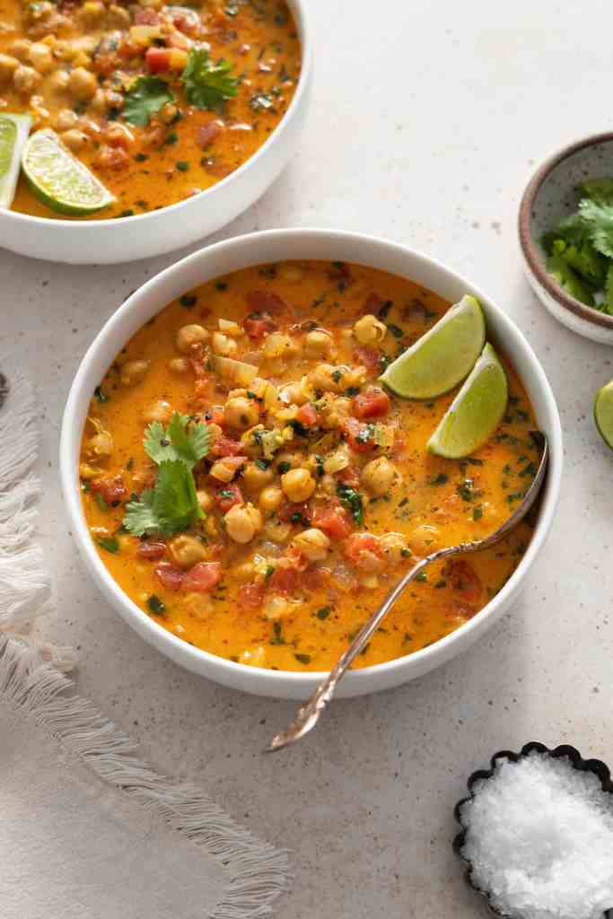 A bowl of creamy chickpea curry soup topped with fresh cilantro and lime wedges, surrounded by salt in a small dish and a textured cloth.