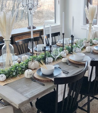 A beautifully styled Thanksgiving tablescape featuring a wood table with black chairs, neutral dishware, white pumpkins, and greenery. Taper candles in black holders and decorative elements like pampas grass add elegance.