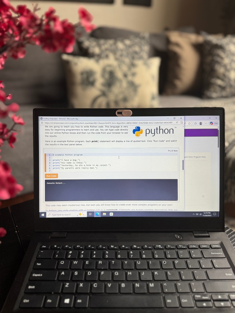 A laptop displaying Python coding instructions and examples on the screen, with a decorative pink flower arrangement in the foreground and a cozy living room background.